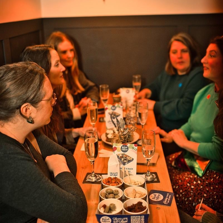 Group of 6 women sitting around a table with glasses of wine and snacks, engaged in conversation.
