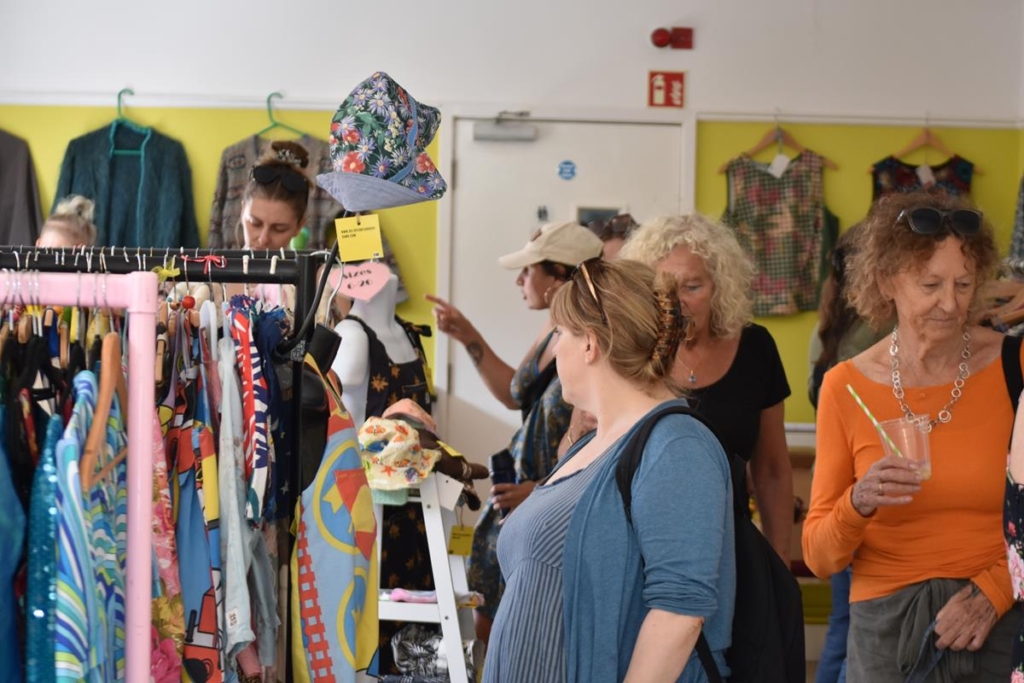 Customers at an indoor clothes market, with rails of brightly coloured clothes.