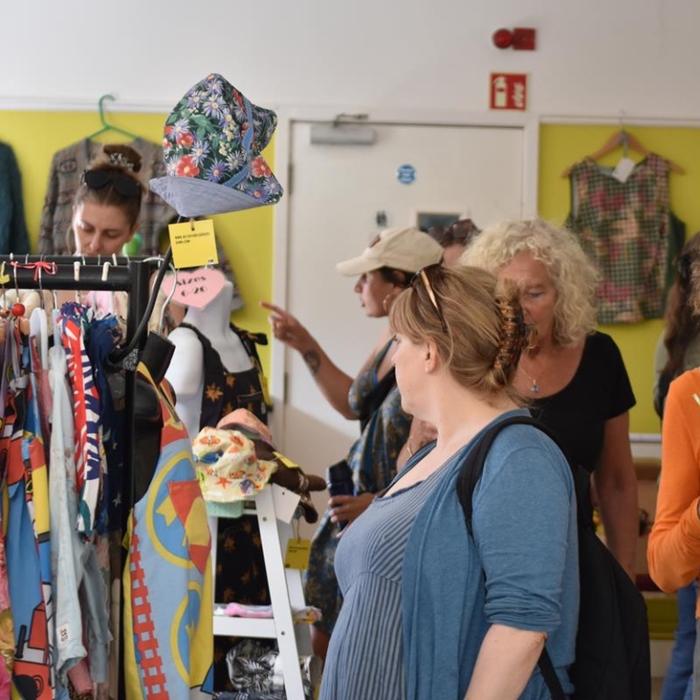 Customers at an indoor clothes market, with rails of brightly coloured clothes.