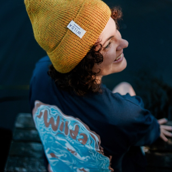 Model wearing a Wilda-branded knitted beanie hat in mustard, and a navy t-shirt with a Wilda graphic featuring swimmers in a lake.